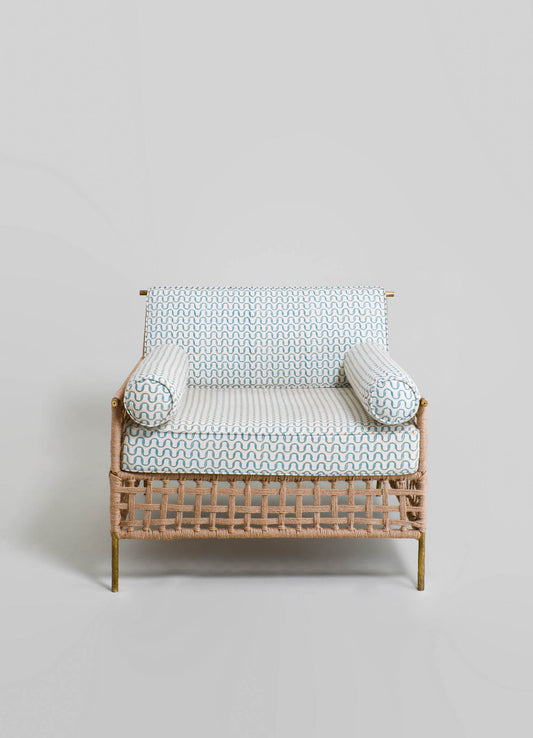 Rattan loveseat with a woven frame, featuring patterned fabric cushions in blue and white geometric motifs, with cylindrical bolster pillows on each side. The design combines rustic and contemporary styles with brass accents.
