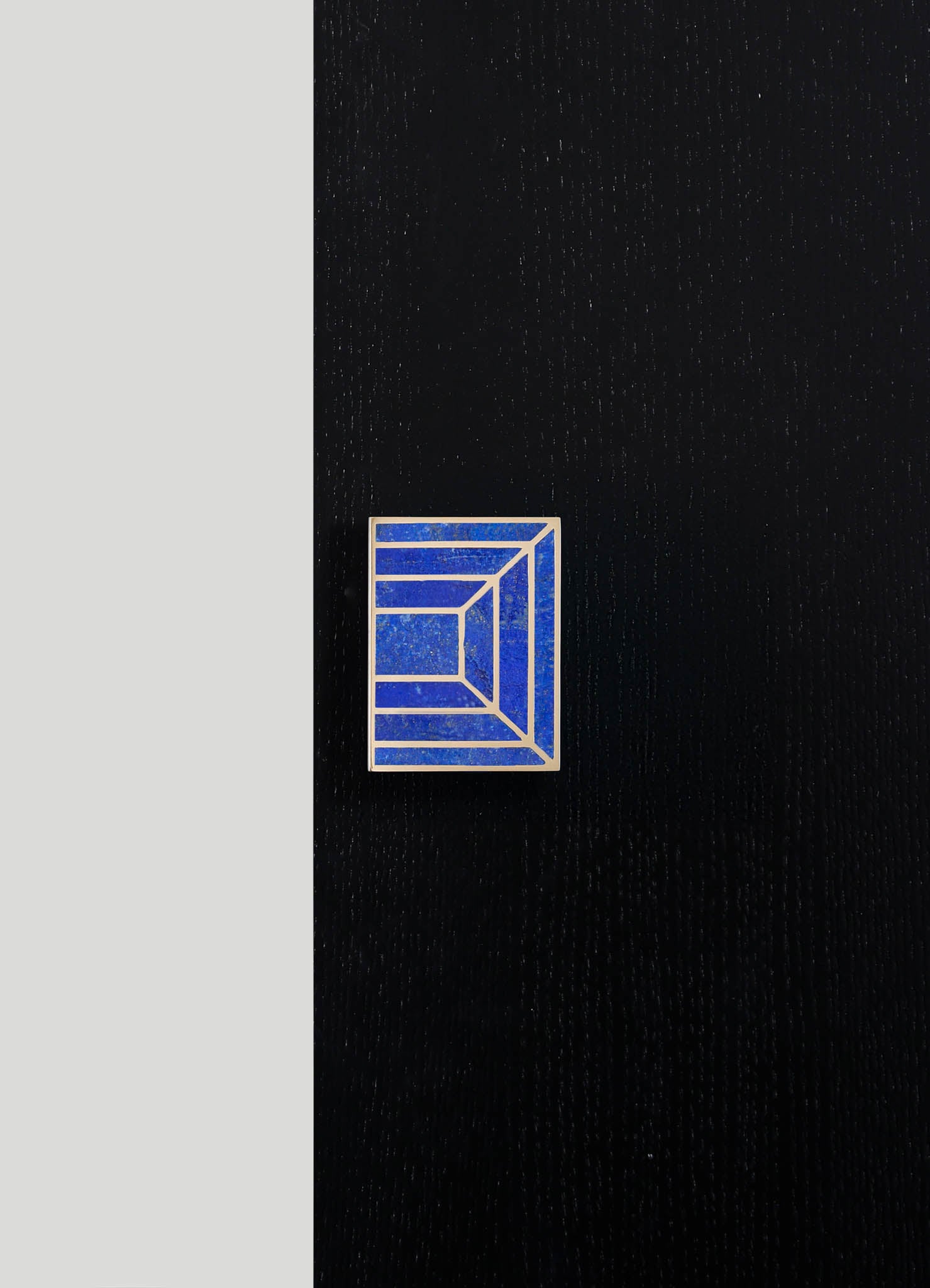 A rectangular lapel pin with geometric lines and a blue textured surface set against black fabric. The pin features silver or metallic borders creating a layered, linear pattern.