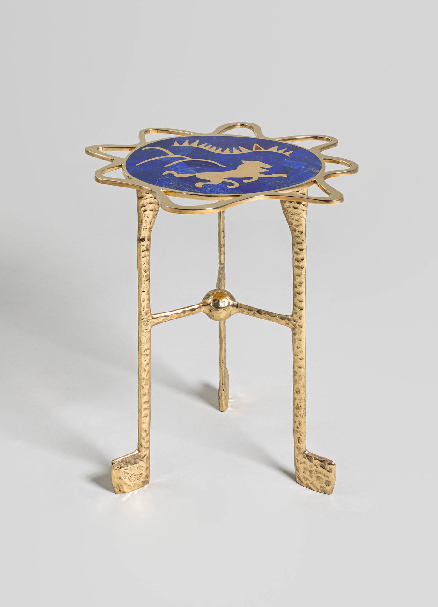 A small side table with a gold, hammered metal leg structure and a round top featuring a blue background with a golden lion motif. The style combines handcrafted metalwork with decorative motifs, creating an elegant and eclectic look.