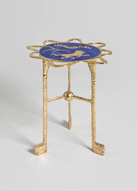 A small side table with a gold, hammered metal leg structure and a round top featuring a blue background with a golden lion motif. The style combines handcrafted metalwork with decorative motifs, creating an elegant and eclectic look.