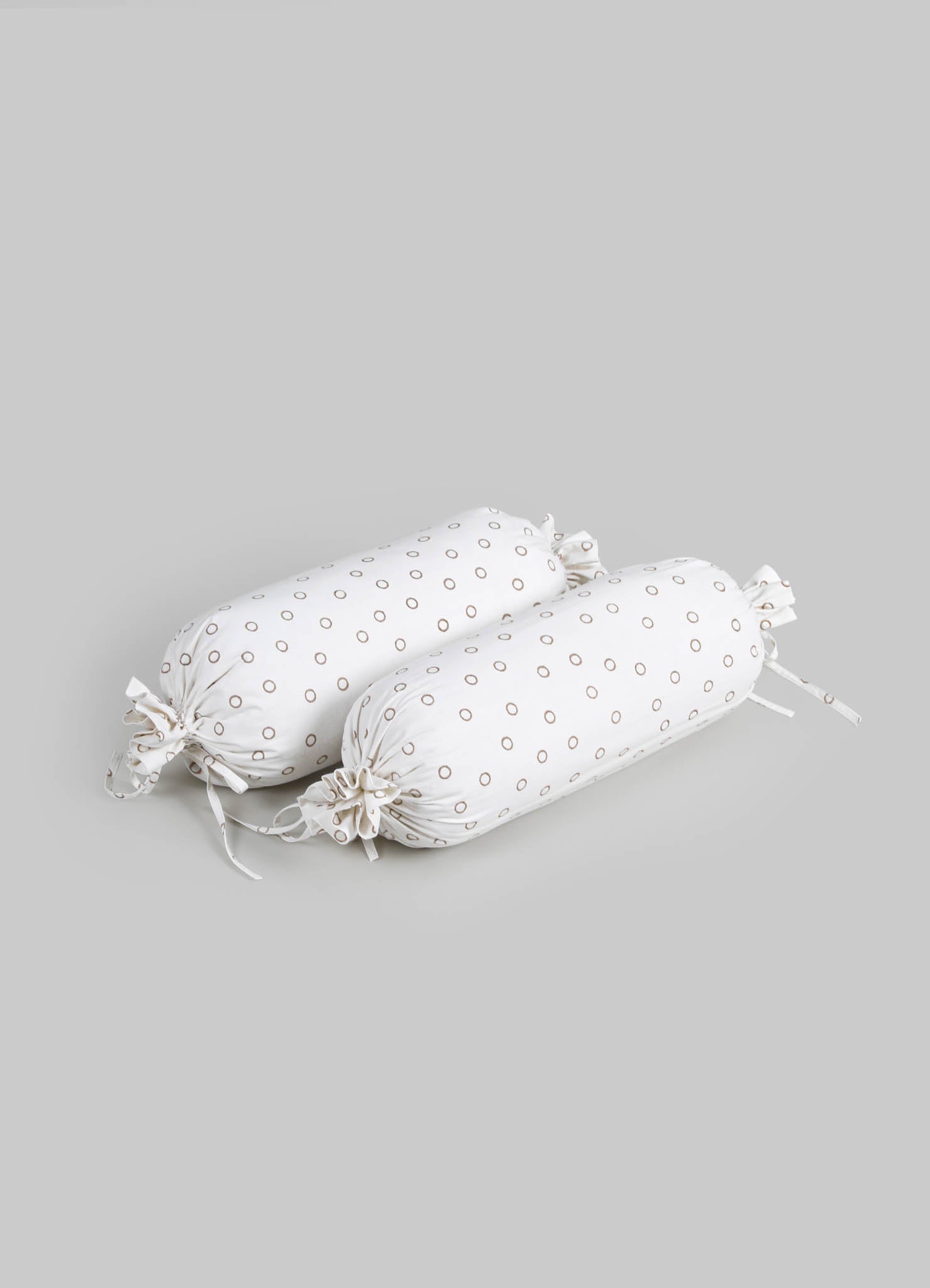 Pair of cylindrical fabric seed pillows with white cotton material and small brown circular motifs, finished with drawstring closures.