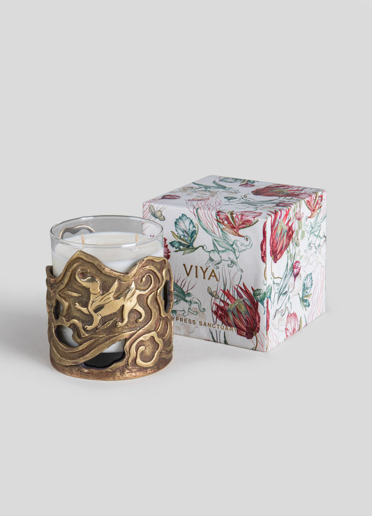 A white scented candle in a decorative brass holder with an intricate, organic motif, accompanied by a floral-illustrated gift box with the label "VIYA" and "Cypress Sanctuary."