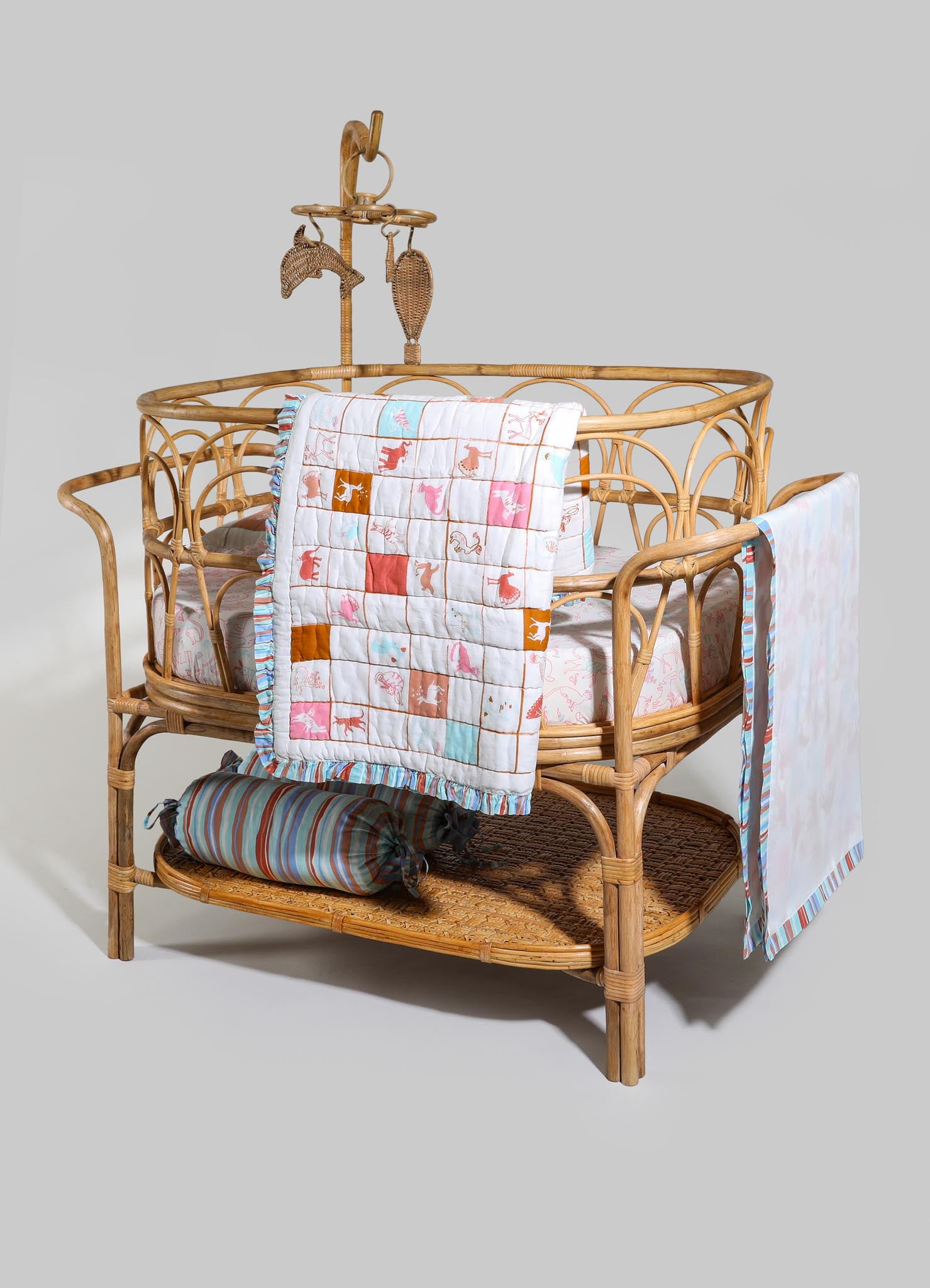 Wicker baby crib with rounded frame and intricate woven design, adorned with pastel-colored textile accessories including a quilt with animal motifs, striped pillow, and fabric drapes, set against a neutral background.