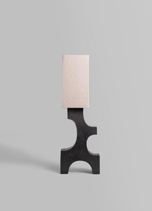 A modern table lamp with a black textured base featuring cut-out geometric shapes and a rectangular fabric lampshade in soft beige, set against a neutral background.