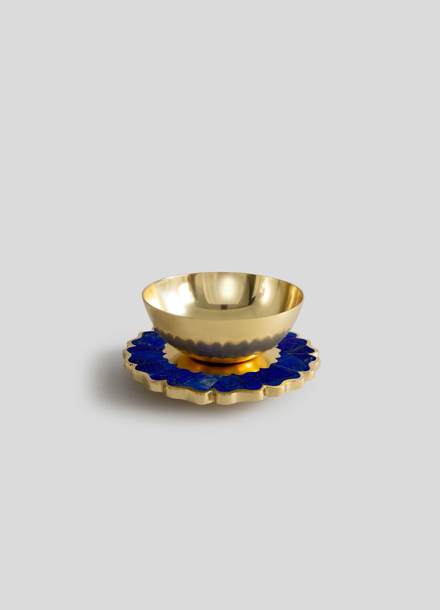 A gold-toned metal bowl with a polished finish, placed on a decorative blue and gold ceramic coaster with a scalloped edge design.