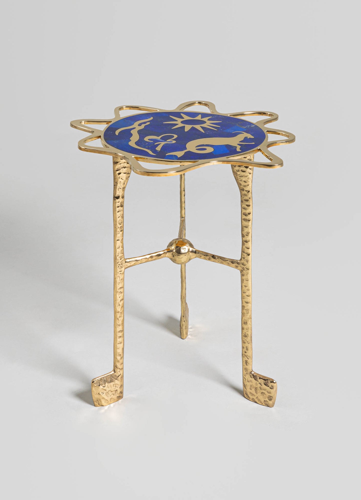 A decorative side table with a brass finish and hammered texture, featuring a circular top with a blue inlay in a sun motif. The table has four slender legs with a handcrafted appearance, combining modern and artisanal design elements.