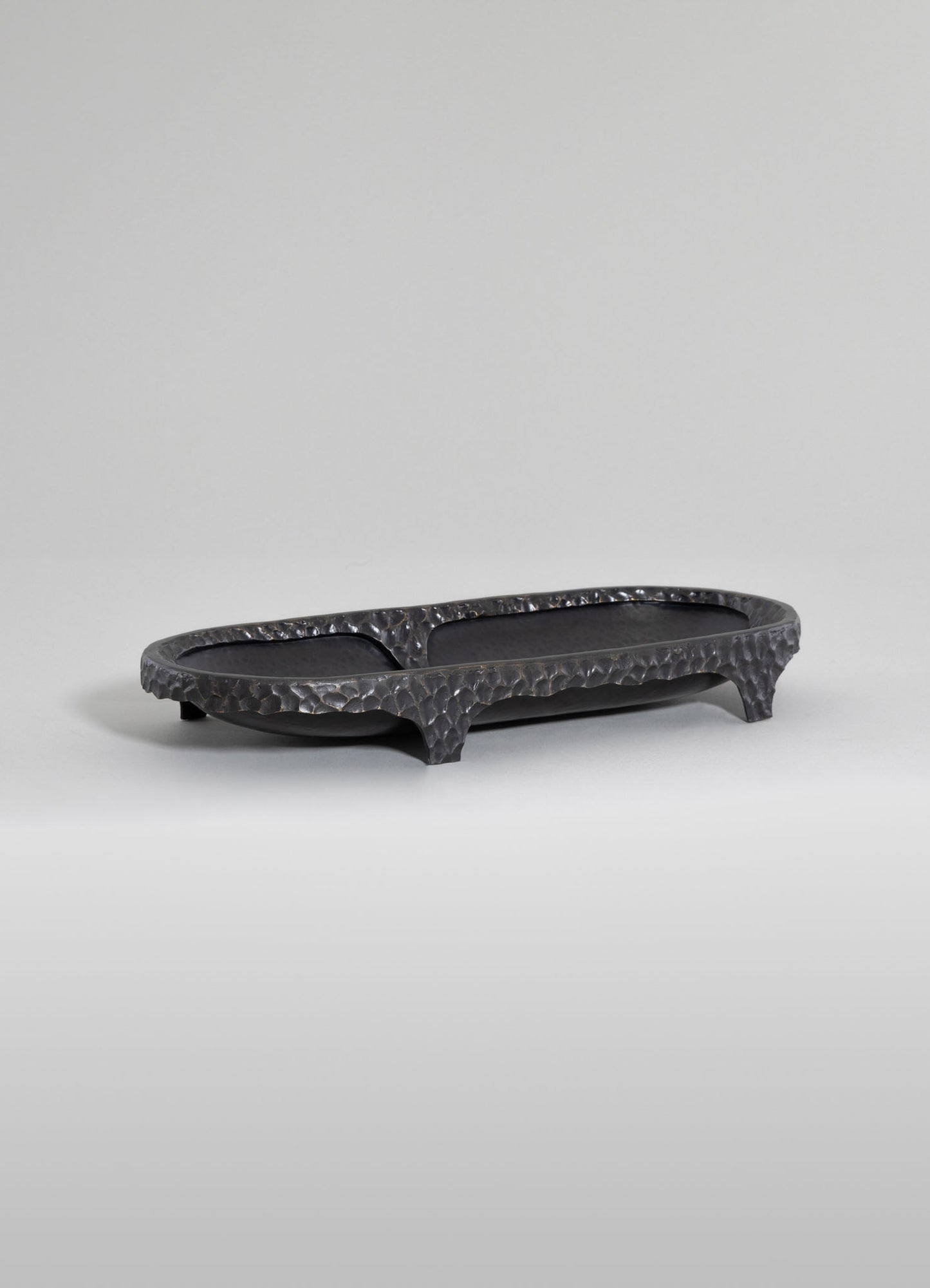 A black hammered metal tray with an elongated oval shape, high edges, and three short legs, featuring a textured and reflective surface.
