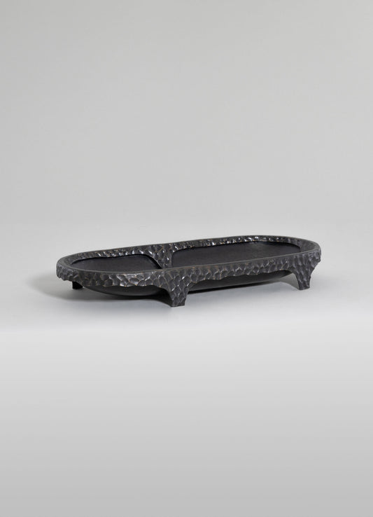 A black hammered metal tray with an elongated oval shape, high edges, and three short legs, featuring a textured and reflective surface.