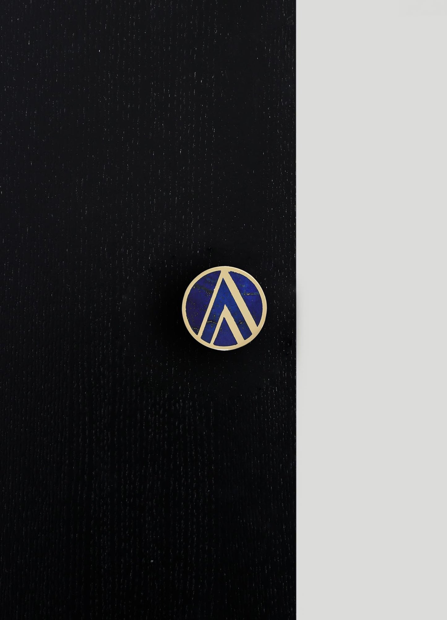 A round lapel pin with a geometric design featuring blue enamel and gold-tone metal borders, set against a textured black background.