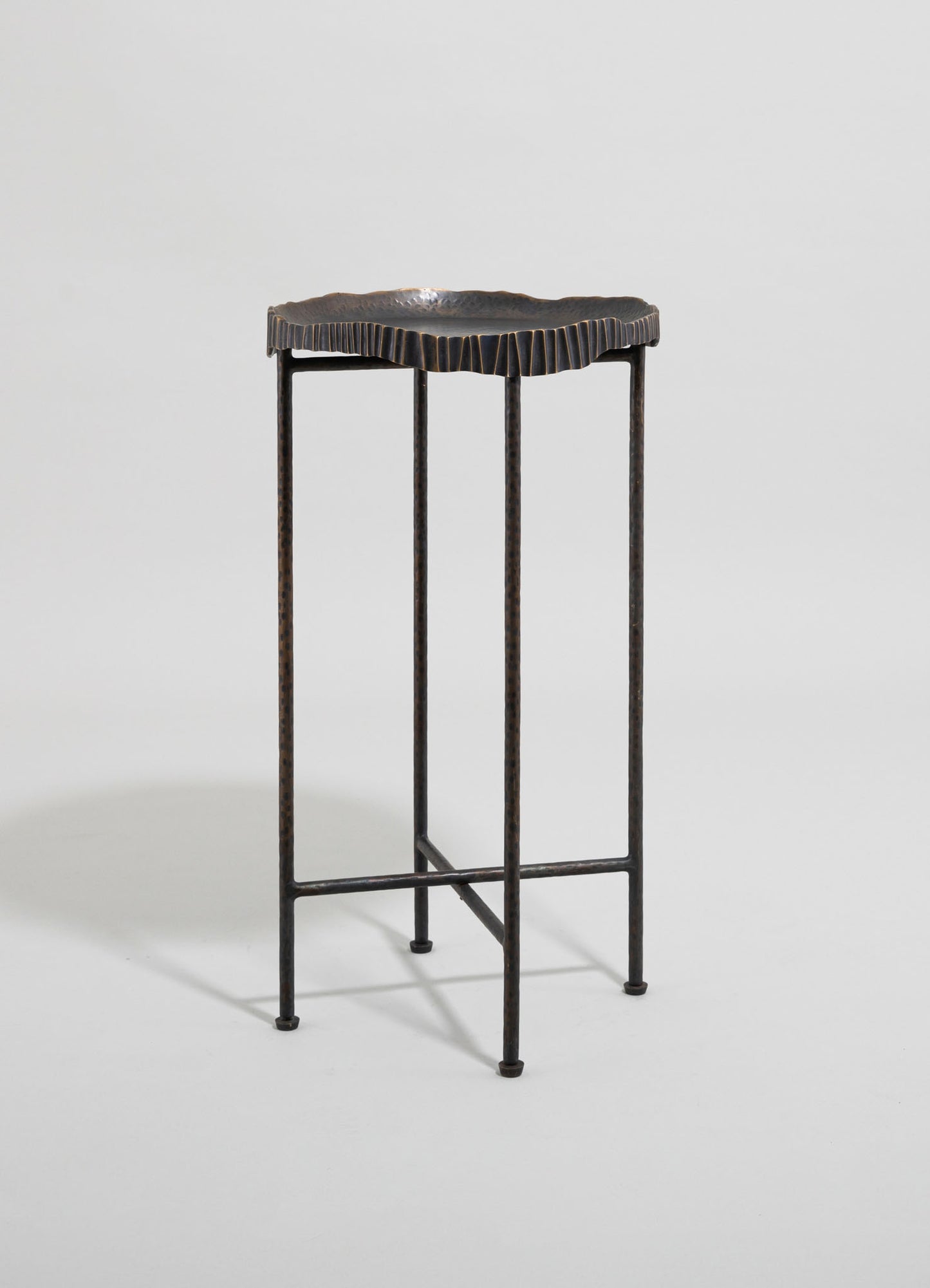 A decorative metal stand with a hammered finish and scalloped edge on the top, featuring a dark, oxidized finish and slender, textured legs.