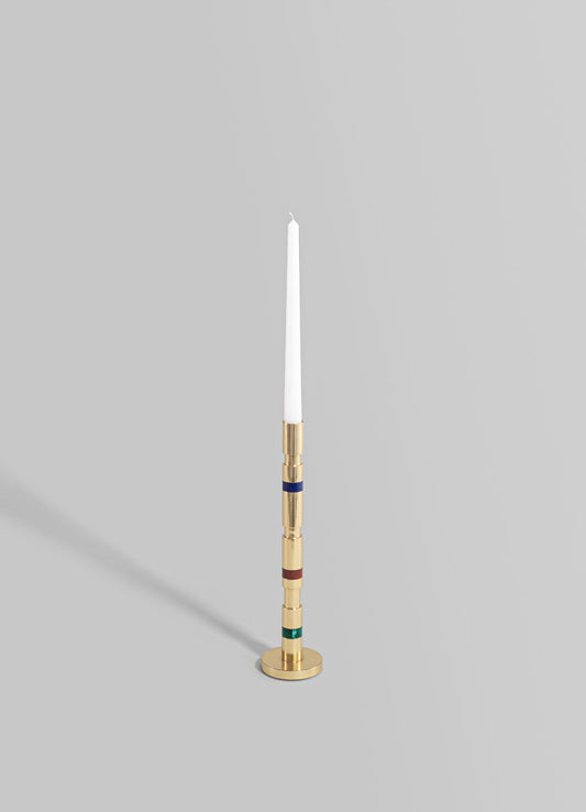 A tall taper candle with a white wax finish in a modern brass candlestick holder featuring multicolored geometric bands, set against a plain gray background.