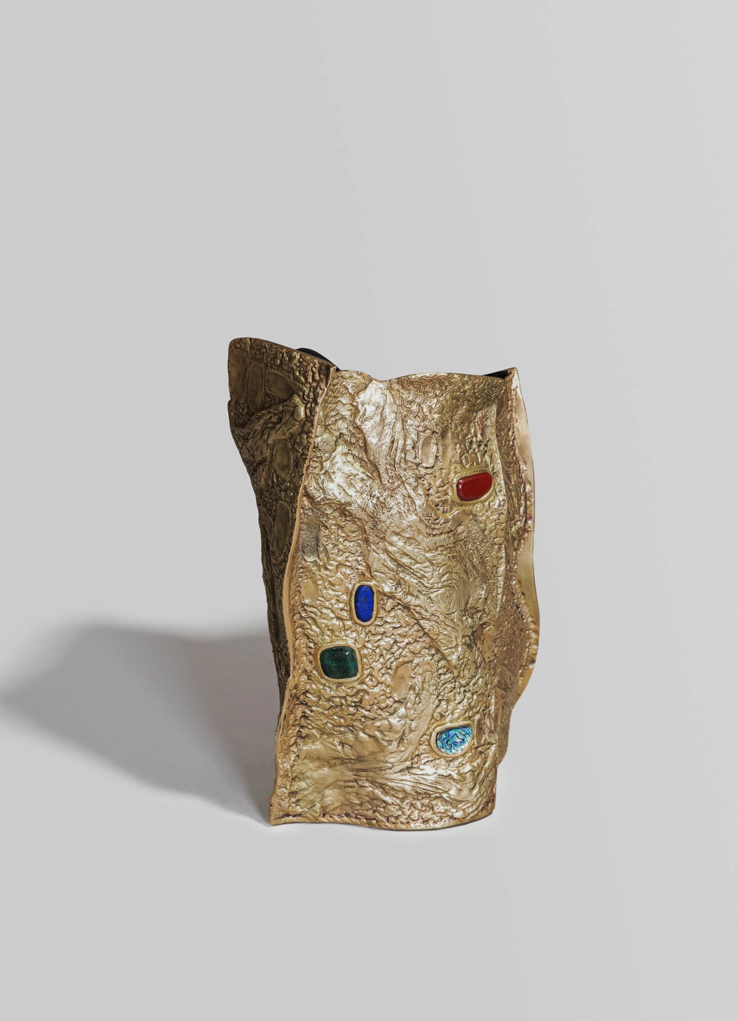 A sculptural vase with a textured gold finish and embedded colorful stones in red, blue, green, and iridescent blue, featuring an organic, irregular shape.