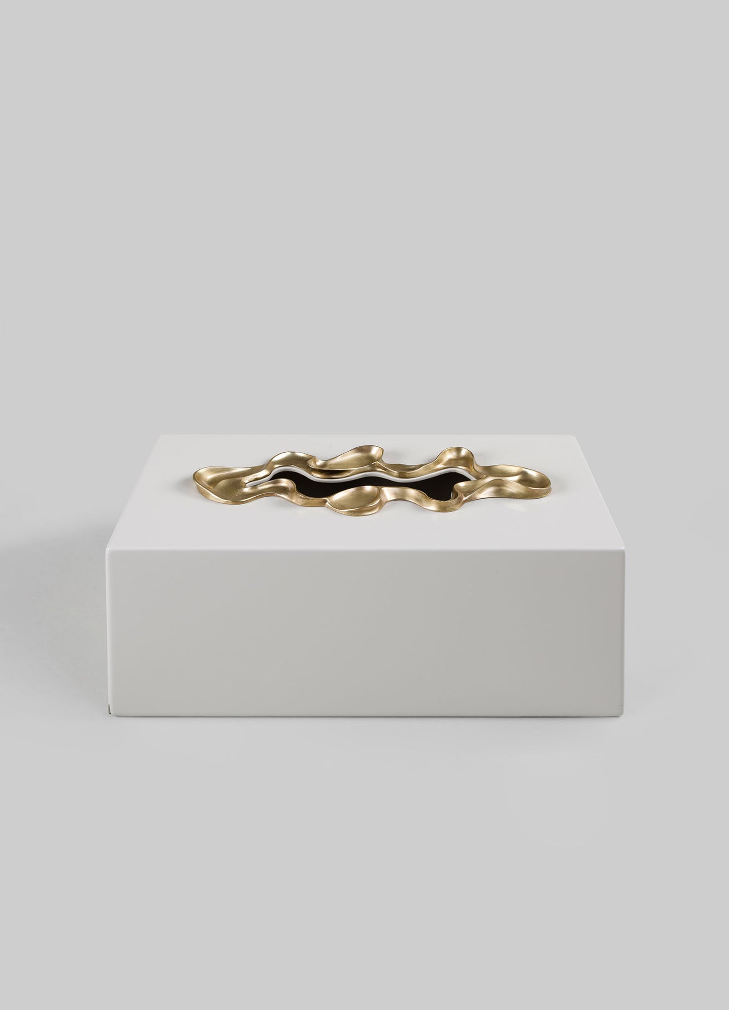 A decorative brass tray with an irregular, wavy edge and a polished finish, placed on a white rectangular box against a plain gray background.