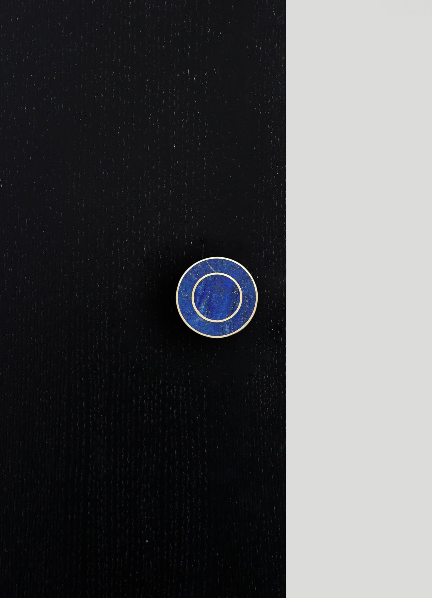 A circular lapel pin made of blue lapis lazuli with gold-tone metal framing, placed on a black textured surface with a contrasting light gray background.