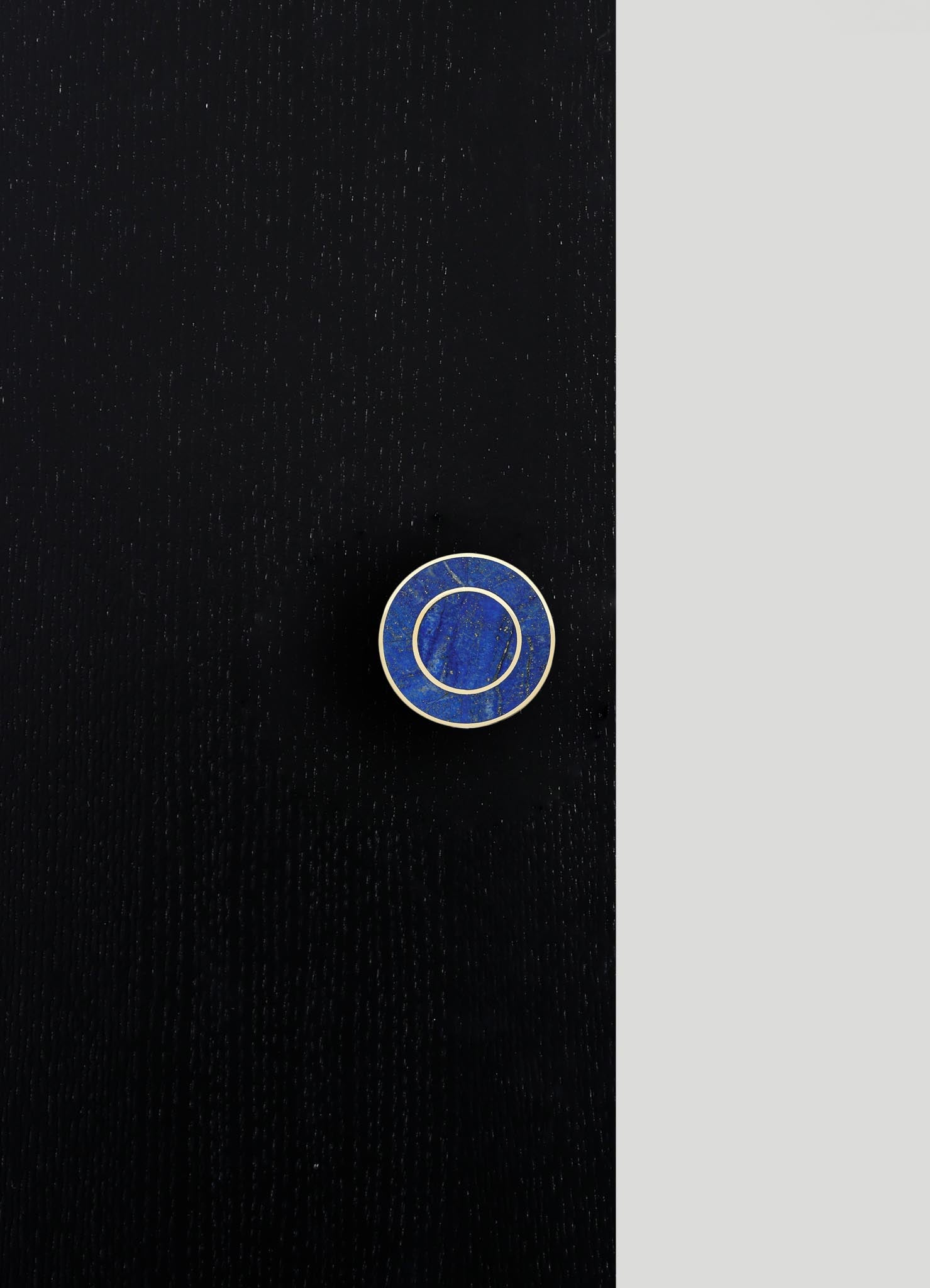 A circular lapel pin made of blue lapis lazuli with gold-tone metal framing, placed on a black textured surface with a contrasting light gray background.