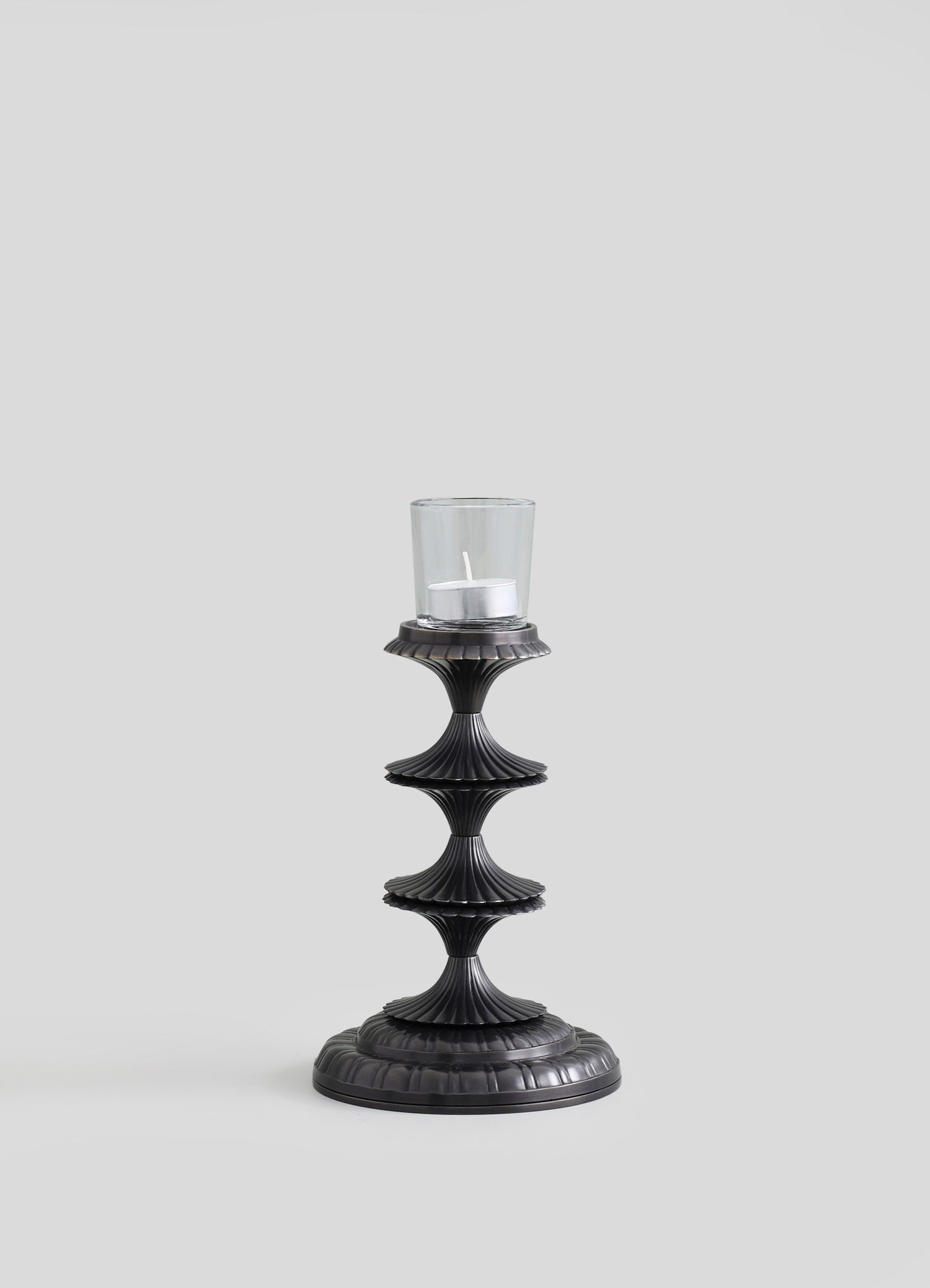A black metal candle holder with a fluted, tiered design and a round base, holding a glass hurricane with a white pillar candle inside.