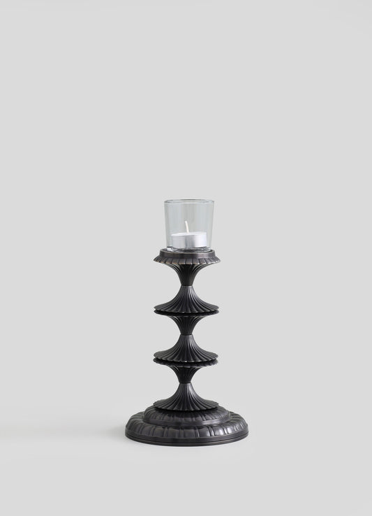 A black metal candle holder with a fluted, tiered design and a round base, holding a glass hurricane with a white pillar candle inside.