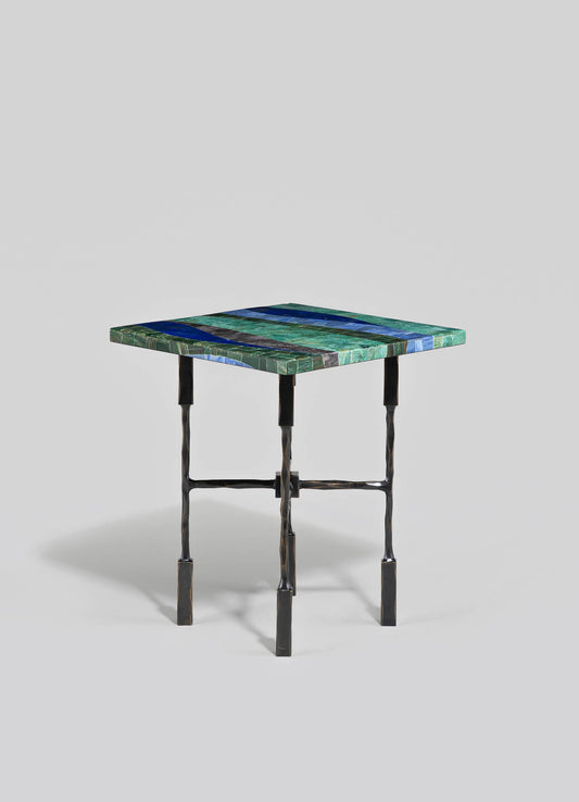 A small table with a rectangular top made of multicolored mosaic tiles in shades of green, blue, and black, supported by a black, hammered metal frame with four slender, slightly twisted legs interconnected by horizontal supports.
