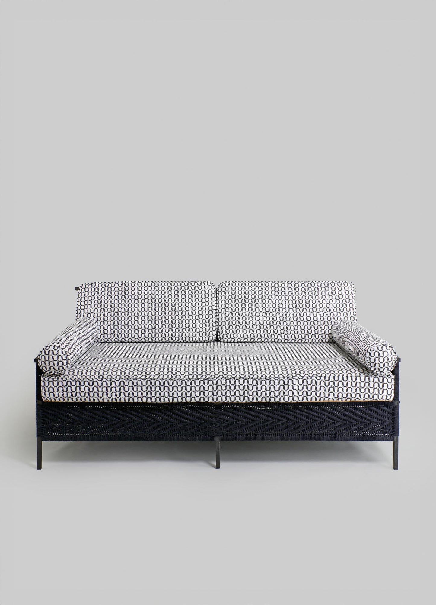A sofa with a woven black base and patterned fabric cushions in black and white geometric designs, paired with matching bolster pillows, set against a plain gray background.