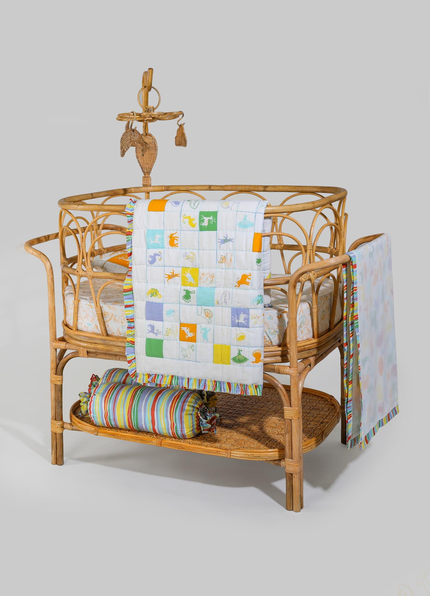 A woven rattan baby crib with a natural finish, featuring a lightweight and rounded frame with open sides, accompanied by colorful, patterned textiles including a quilt with animal motifs, and matching striped fabric accents. A small, woven rattan mobile with hanging decorations is attached to the top of the crib.