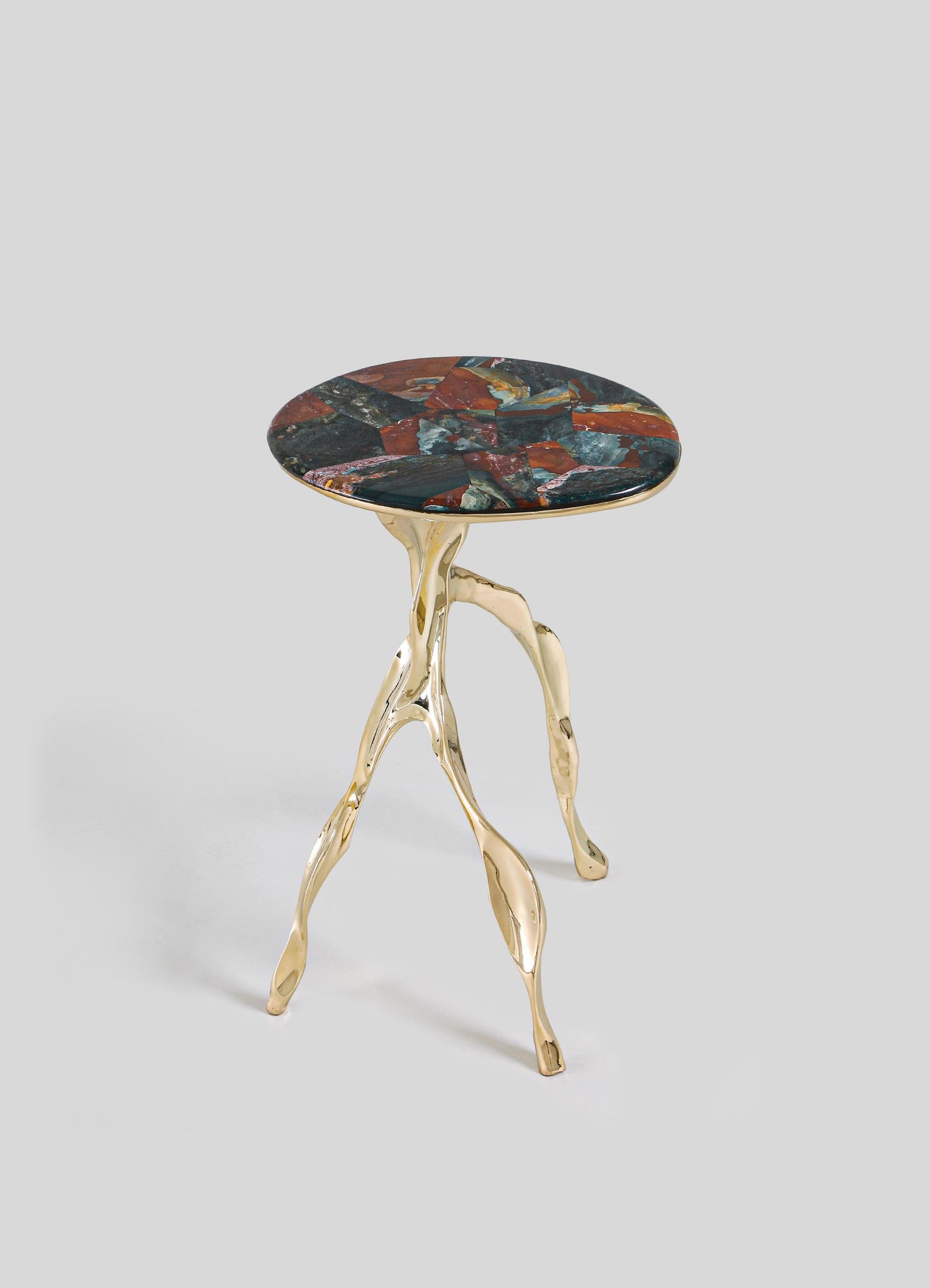 The VIYA Under The Banyan Gold Side Table Large features three gold, branch-like legs inspired by aerial roots and a round, multicolored stone top in red, blue, and dark hues, set against a light gray background.