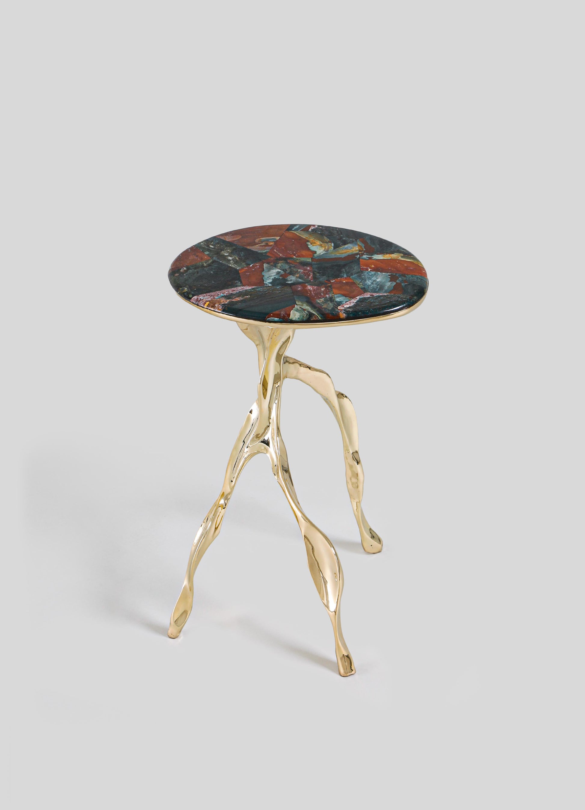 The VIYA Under The Banyan Gold Side Table Large features three gold, branch-like legs inspired by aerial roots and a round, multicolored stone top in red, blue, and dark hues, set against a light gray background.