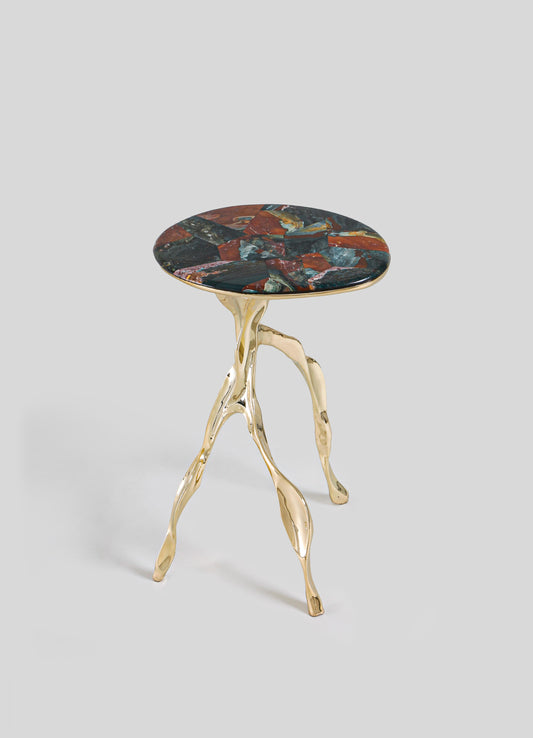 The VIYA Under The Banyan Gold Side Table Large features three gold, branch-like legs inspired by aerial roots and a round, multicolored stone top in red, blue, and dark hues, set against a light gray background.