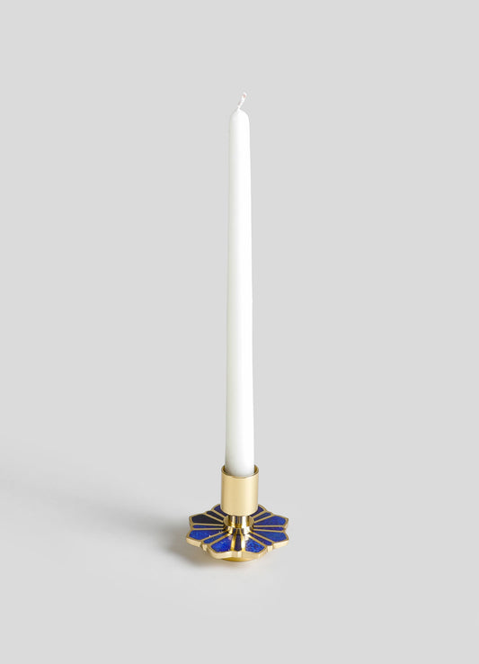 A tall white taper candle in a gold and blue enamel holder with a floral motif design, set against a neutral background.