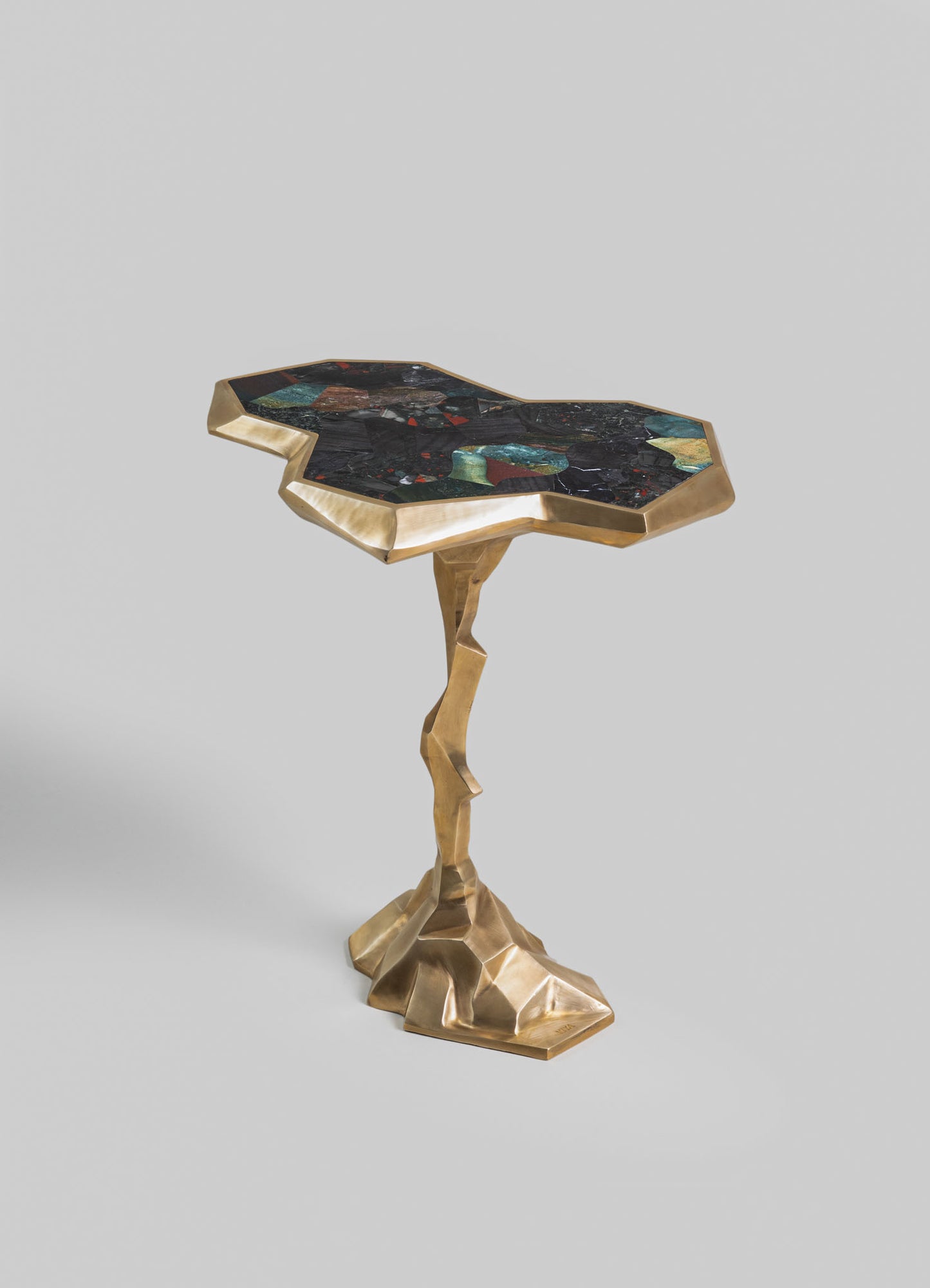A side table with an irregularly shaped top made of multicolored stone in shades of green, red, and black, bordered by a gold-toned metal frame. The base is a sculptural, geometric gold metal structure with an organic, twisted form.