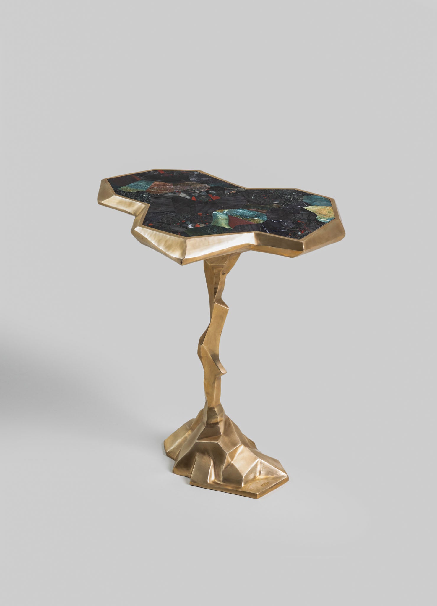 A side table with an irregularly shaped top made of multicolored stone in shades of green, red, and black, bordered by a gold-toned metal frame. The base is a sculptural, geometric gold metal structure with an organic, twisted form.
