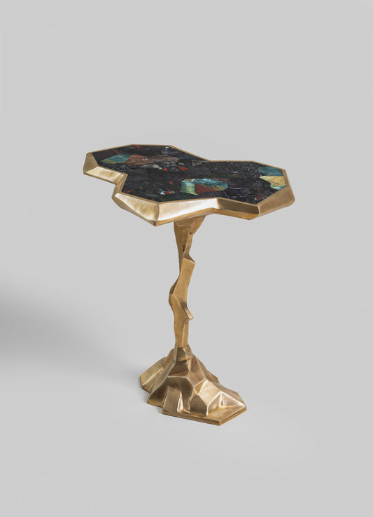 A side table with an irregularly shaped top made of multicolored stone in shades of green, red, and black, bordered by a gold-toned metal frame. The base is a sculptural, geometric gold metal structure with an organic, twisted form.