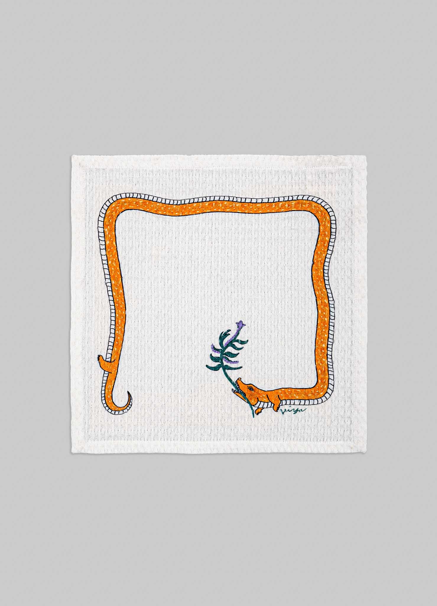 A white cotton napkin featuring an embroidered design of an orange snake with black and white details, coiled around a small purple and green plant motif. The embroidery creates a playful and artistic pattern on the fabric.