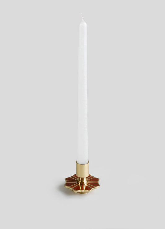 A tall white taper candle with a textured surface, held in a decorative candle holder featuring a gold base and a red geometric pattern resembling petals or a flower shape.
