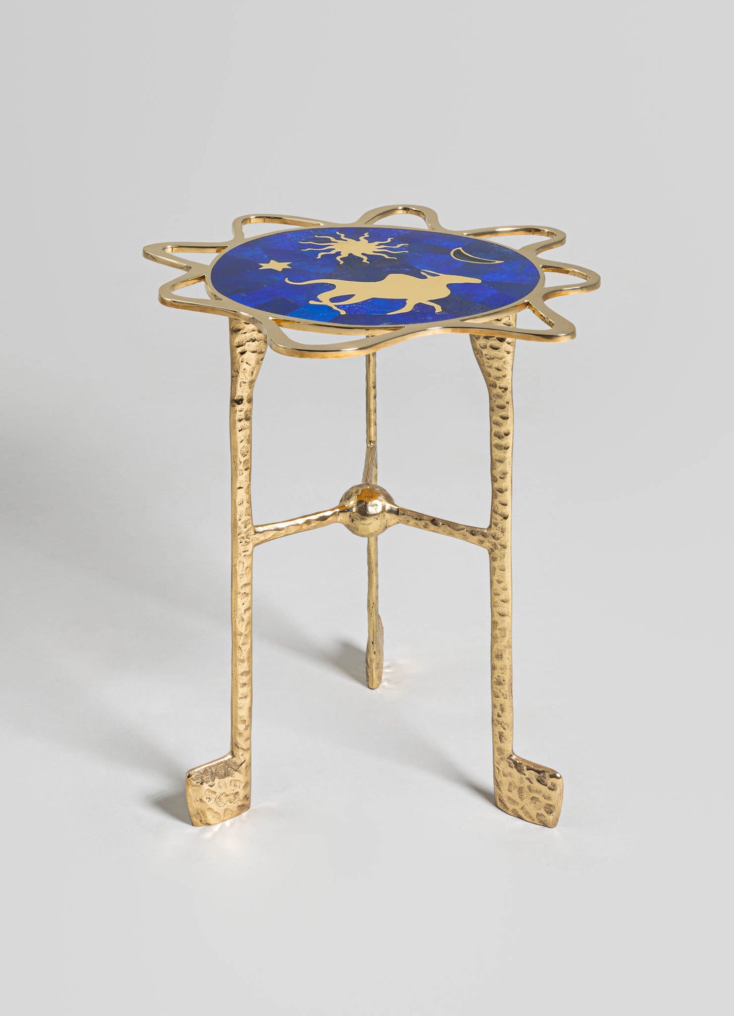 A small side table with a circular top featuring a celestial motif in gold against a deep blue background, framed by an irregular gold metal border. The table has three textured, hammered gold metal legs with a rustic finish.
