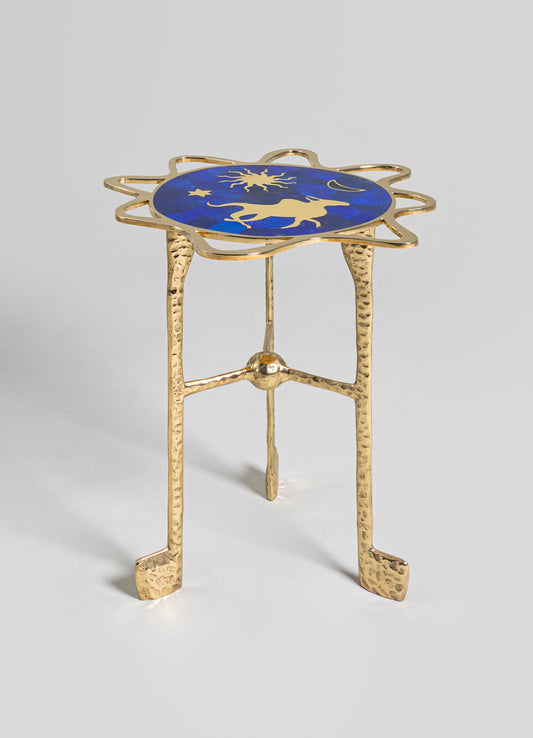 A small side table with a circular top featuring a celestial motif in gold against a deep blue background, framed by an irregular gold metal border. The table has three textured, hammered gold metal legs with a rustic finish.