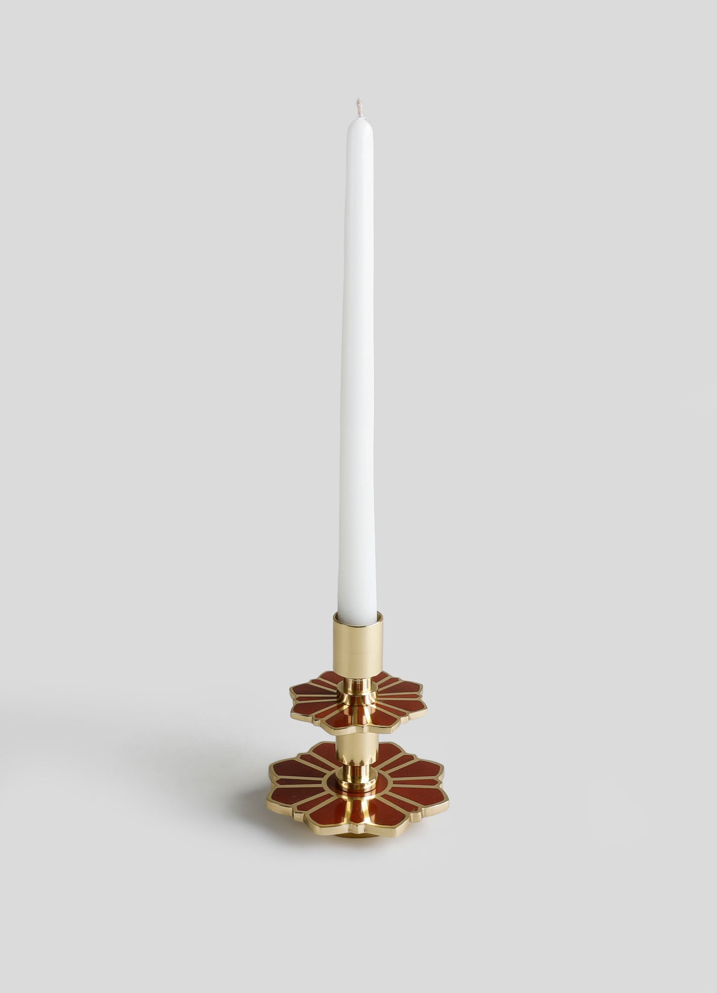 A candlestick holder with a metallic gold finish and red accents in a geometric floral motif, holding a tall white taper candle.