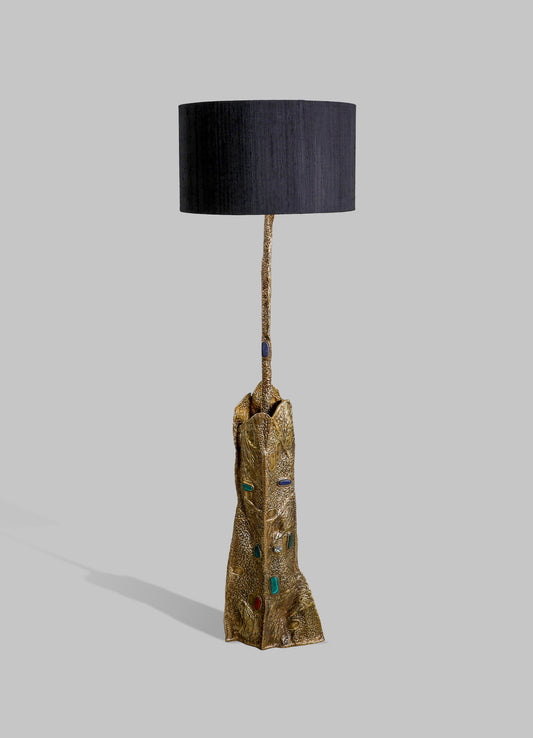 A floor lamp featuring a carved, textured gold-tone base adorned with multicolored accents and a tall, slender stem. The lamp has a large, cylindrical black fabric lampshade with a contemporary style.