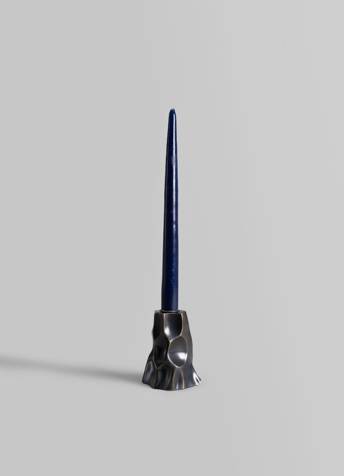 A sculptural candlestick holder with an irregular, textured metal base in a dark, polished finish supporting a tall, slender navy blue candle.