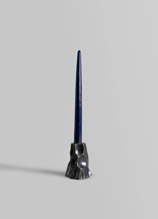 A sculptural candlestick holder with an irregular, textured metal base in a dark, polished finish supporting a tall, slender navy blue candle.