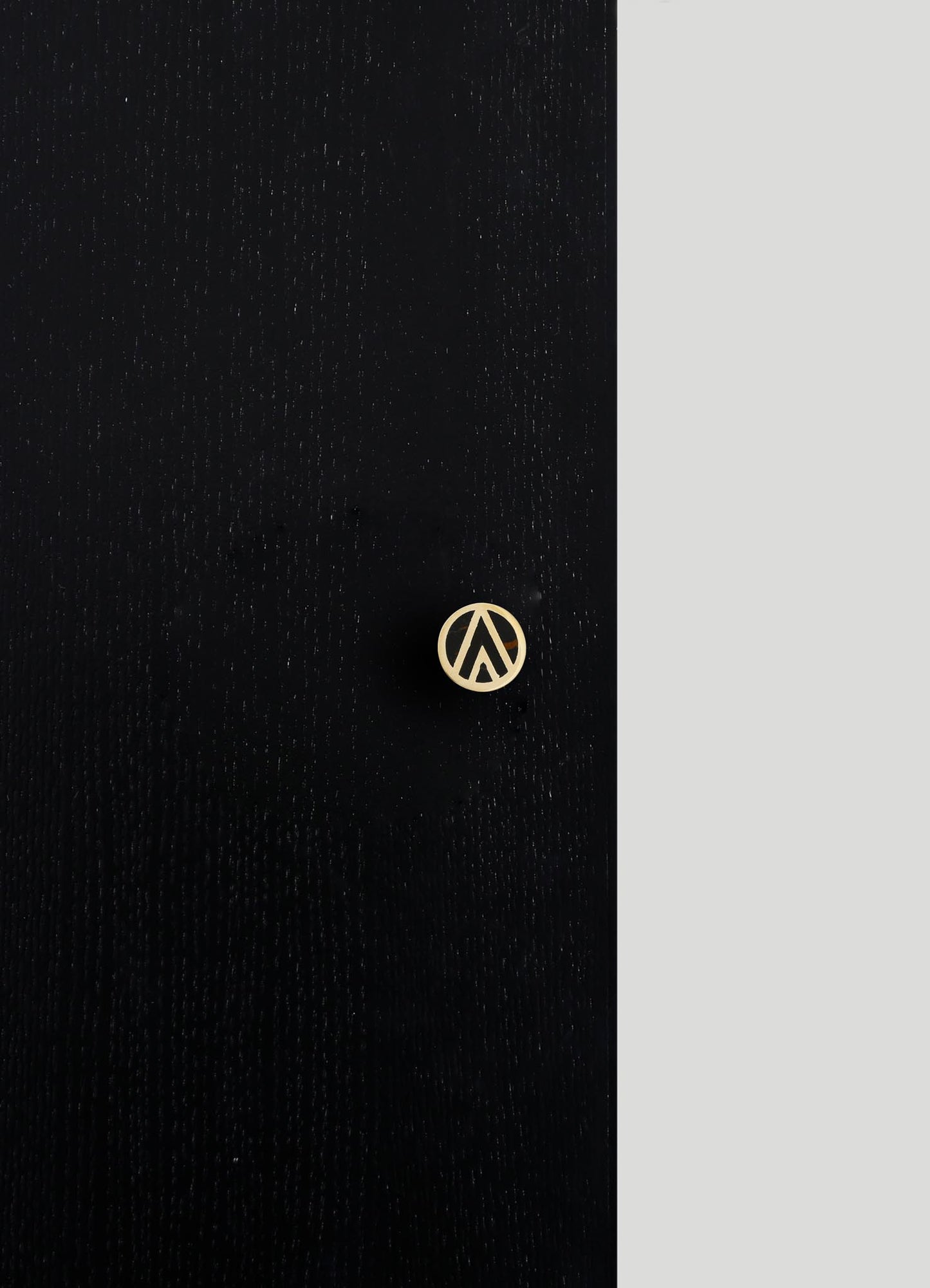 A black wooden surface with a subtle grain texture, featuring a small round brass emblem with a geometric cutout pattern. The background shows a light gray wall.