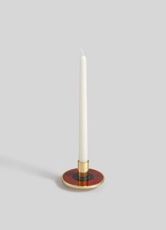 A tall white taper candle with a wicks at the top, placed in a round candle holder featuring a brass base and a red and black patterned surface.
