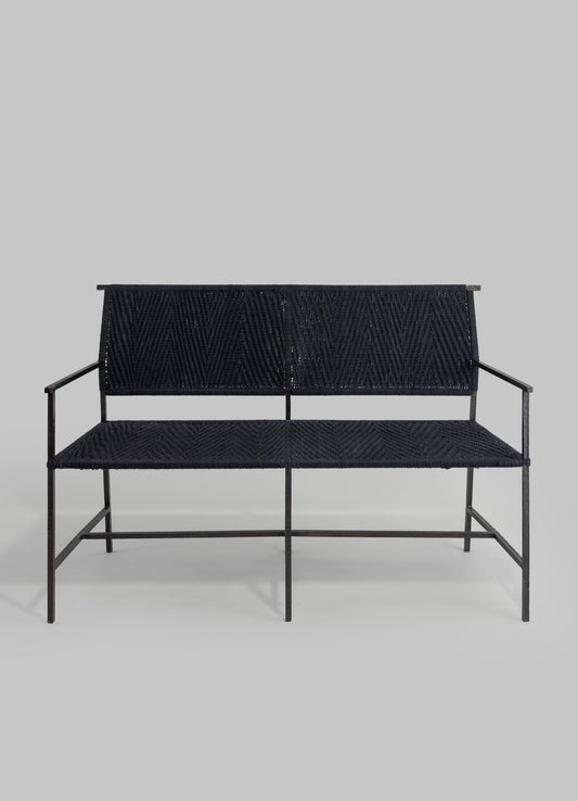 A black metal-framed bench with woven rattan or wicker seat and backrest in a herringbone pattern, set against a plain gray background.