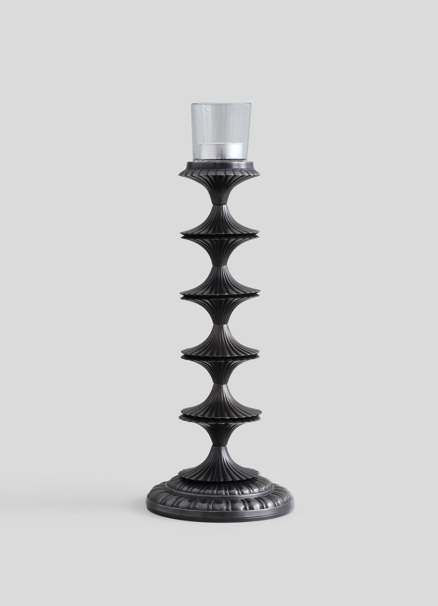 A tall candle holder with a black, textured finish features a series of fluted, hourglass-shaped sections and a round base, designed to hold a glass candle with a metal band around the top.