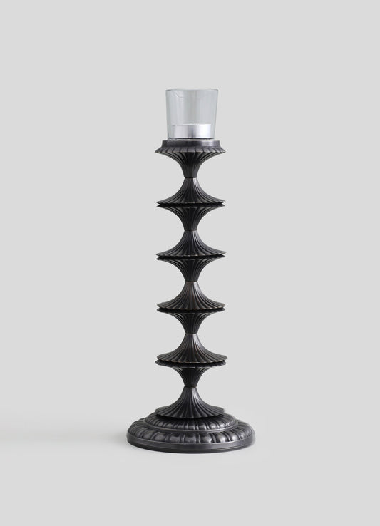 A tall candle holder with a black, textured finish features a series of fluted, hourglass-shaped sections and a round base, designed to hold a glass candle with a metal band around the top.