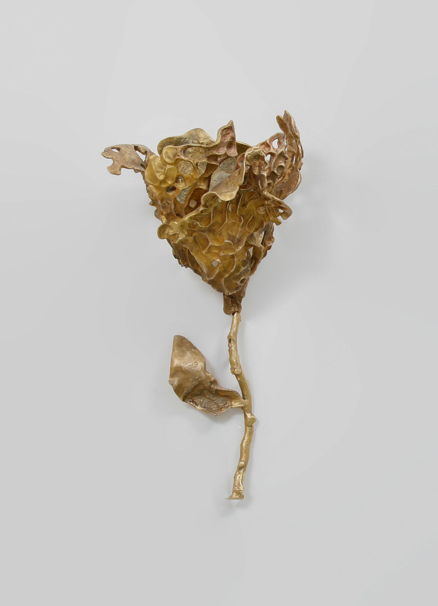 A metallic gold wall sculpture resembling a flower with textured, abstract petals and a long stem. The piece has a contemporary style with organic, sculptural motifs and a polished finish.