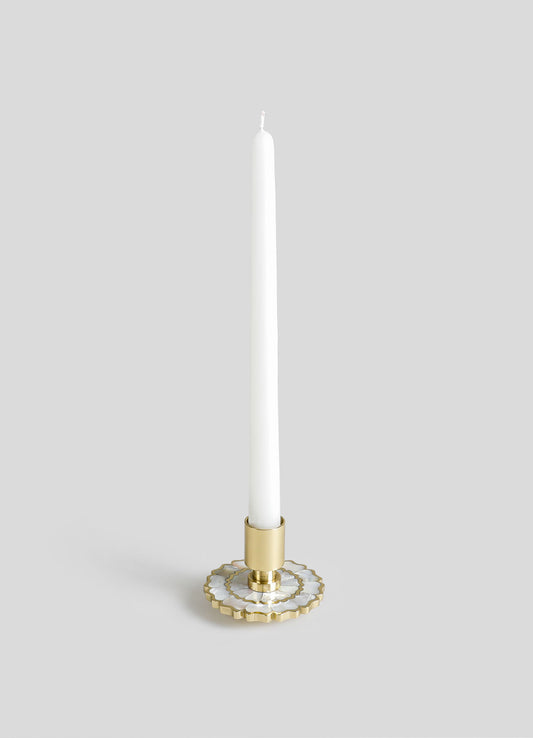 A tall white candle with a slender shape in a decorative brass and white ceramic holder featuring geometric scalloped patterns, set against a neutral background.
