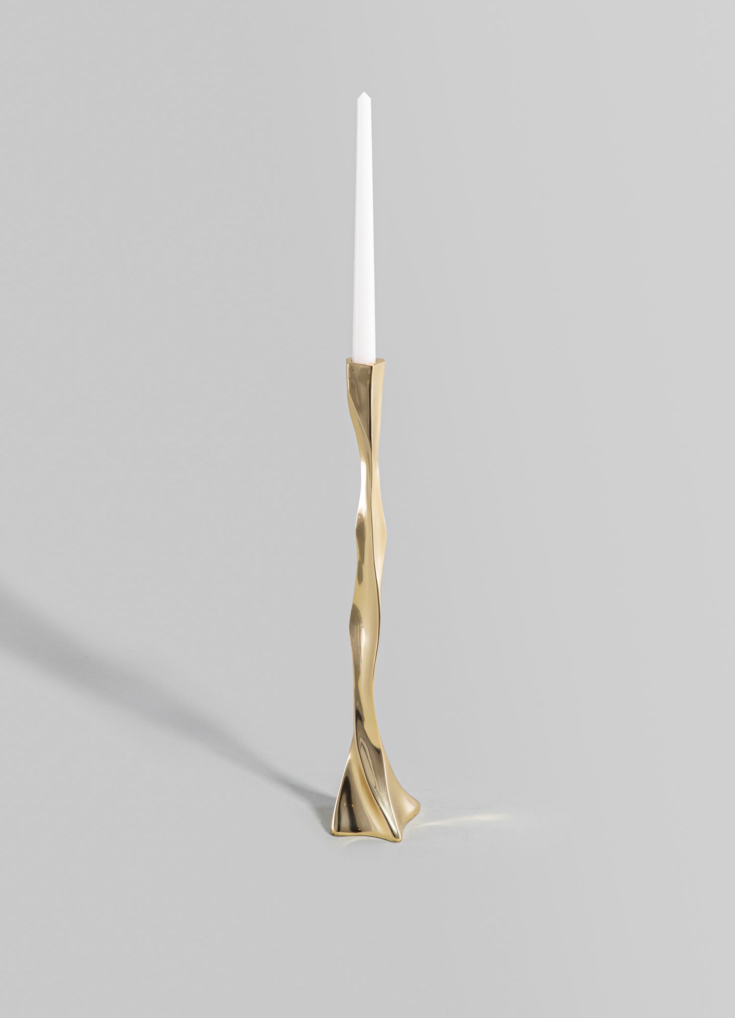 A tall candlestick holder with an irregular, sculptural brass finish and a slim white candle on top, featuring a contemporary and artistic design.