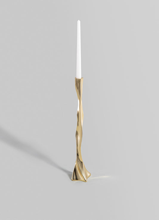 A tall candlestick holder with an irregular, sculptural brass finish and a slim white candle on top, featuring a contemporary and artistic design.