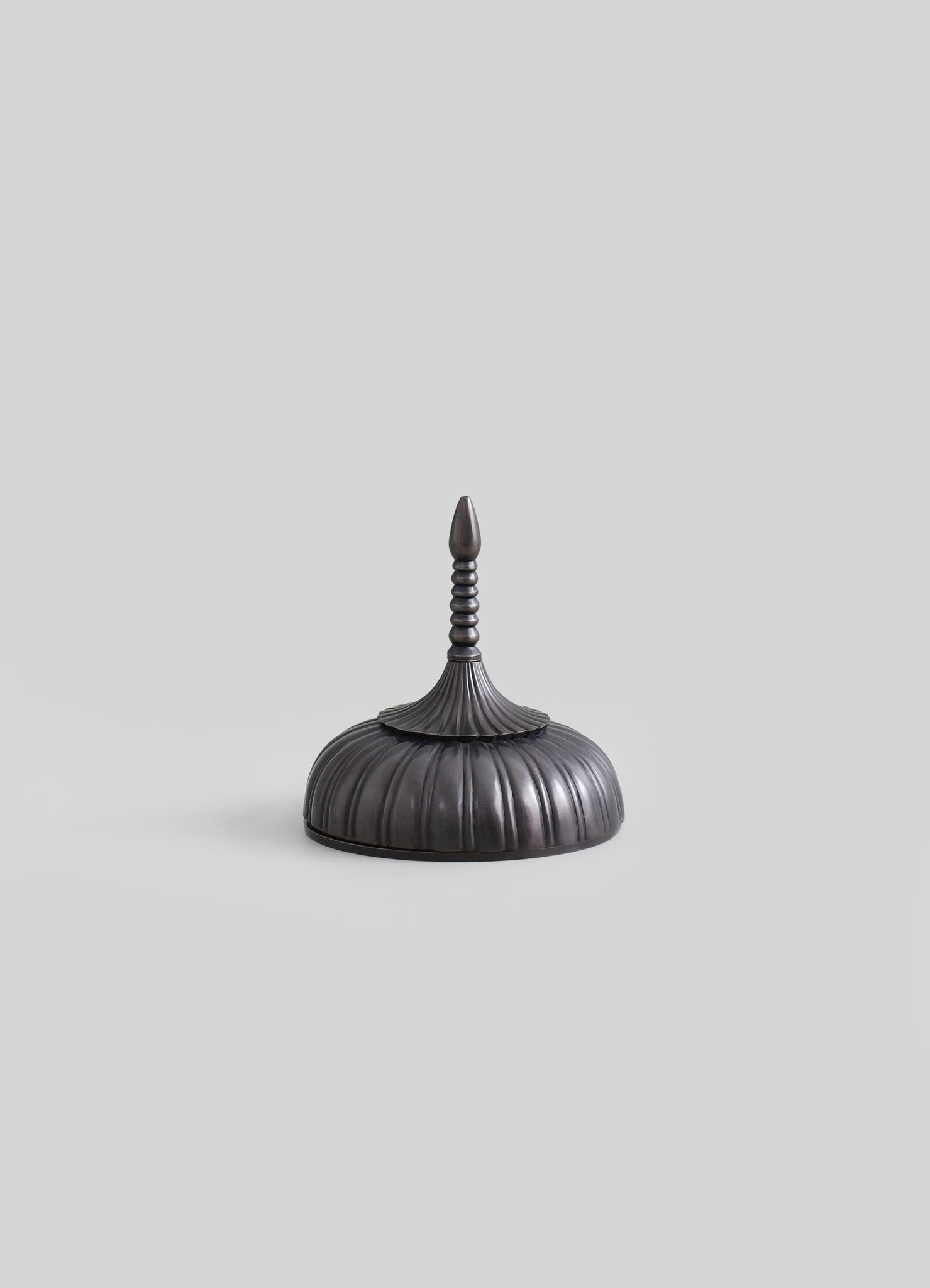 A decorative black metal finial with a rounded, ribbed base, a flared top, and a vertically striped, pointed spire on top, set against a plain light gray background.