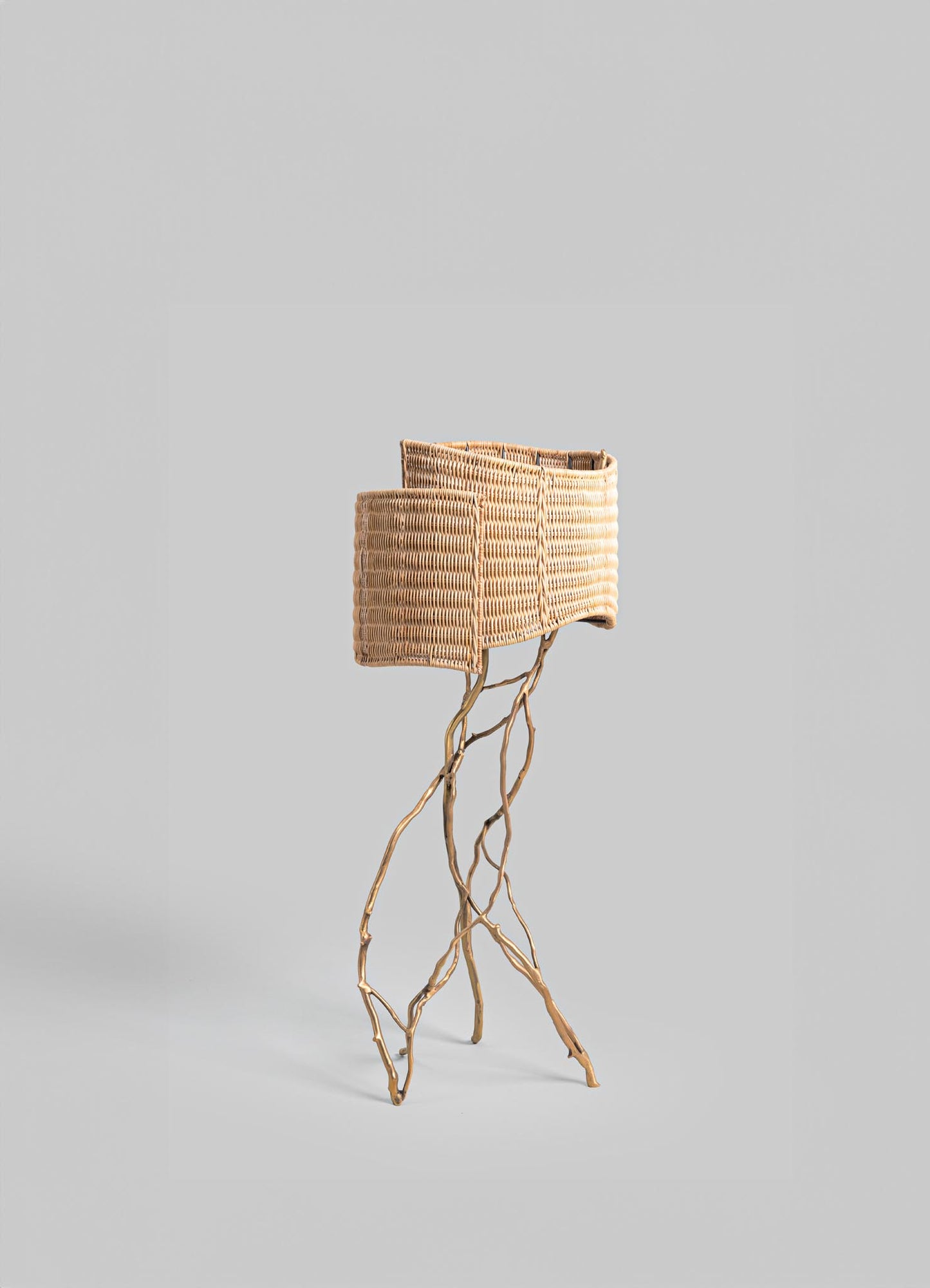 A table lamp with a woven rattan shade and a base made of natural, twisted wood branches, creating a rustic and organic style. The neutral tone of the rattan complements the light wooden stand against a plain background.